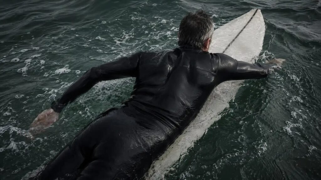 Surfista adulto en prone paddleando, vista cenital, neopreno oscuro, escápulas y tren superior en trabajo activo, agua cantábrica