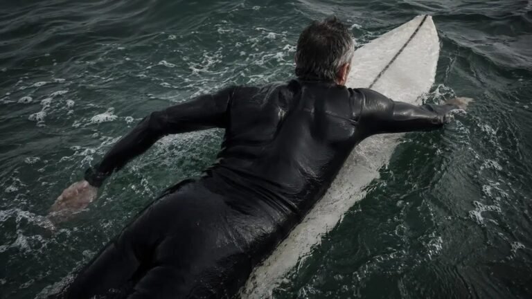 Surfista adulto en prone paddleando, vista cenital, neopreno oscuro, escápulas y tren superior en trabajo activo, agua cantábrica